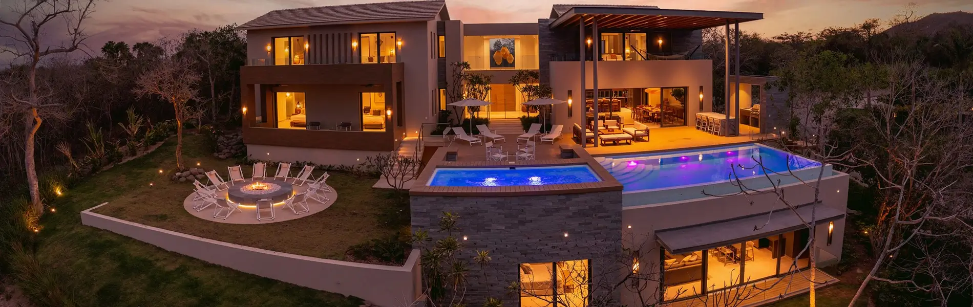 Modern luxury villa with infinity pool and fire pit, illuminated at dusk.