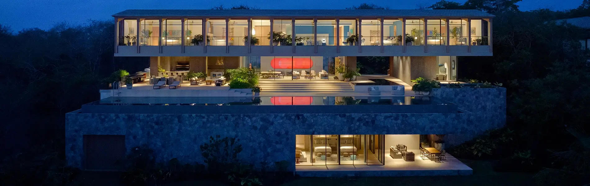 Modern glass-walled villa with infinity pool, illuminated at night